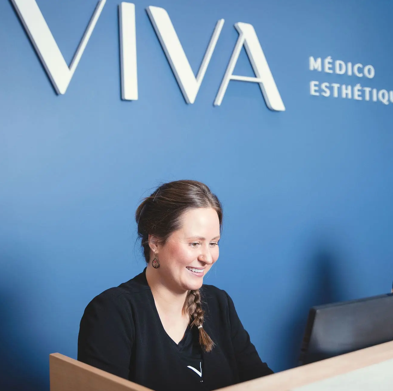 Une réceptionniste souriante avec une tresse brune est assise à la réception devant un mur bleu arborant « VIVA MÉDICO ESTHÉTIQUE », vous souhaitant la bienvenue à la Clinique médico-esthétique Viva.