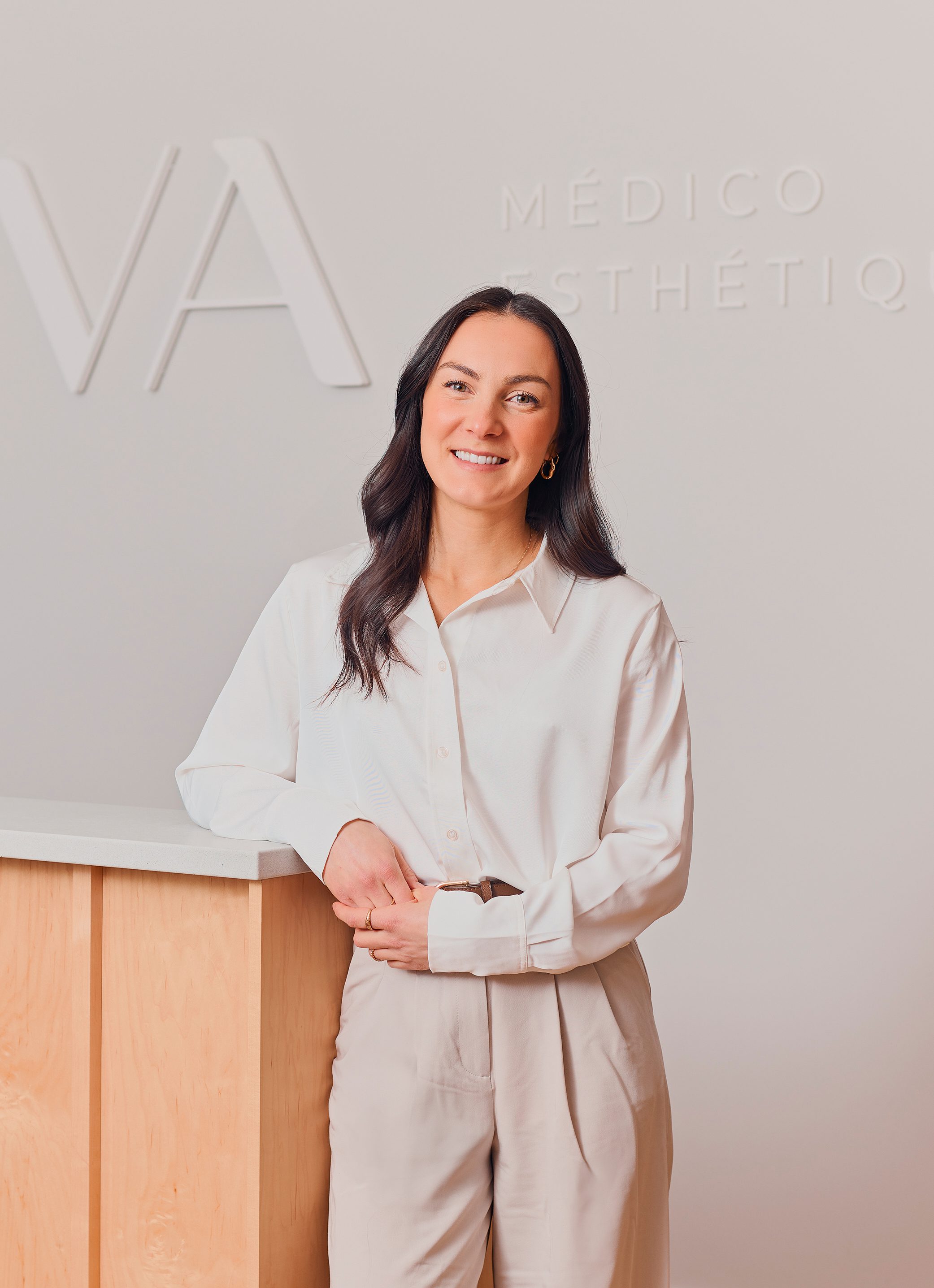 Une femme aux longs cheveux noirs, vêtue d'une chemise blanche boutonnée et d'un pantalon beige, se tient debout, souriante, appuyée contre un comptoir en bois clair de la Clinique médico-esthétique Viva. En arrière-plan, un mur clair avec une partie du texte en français.