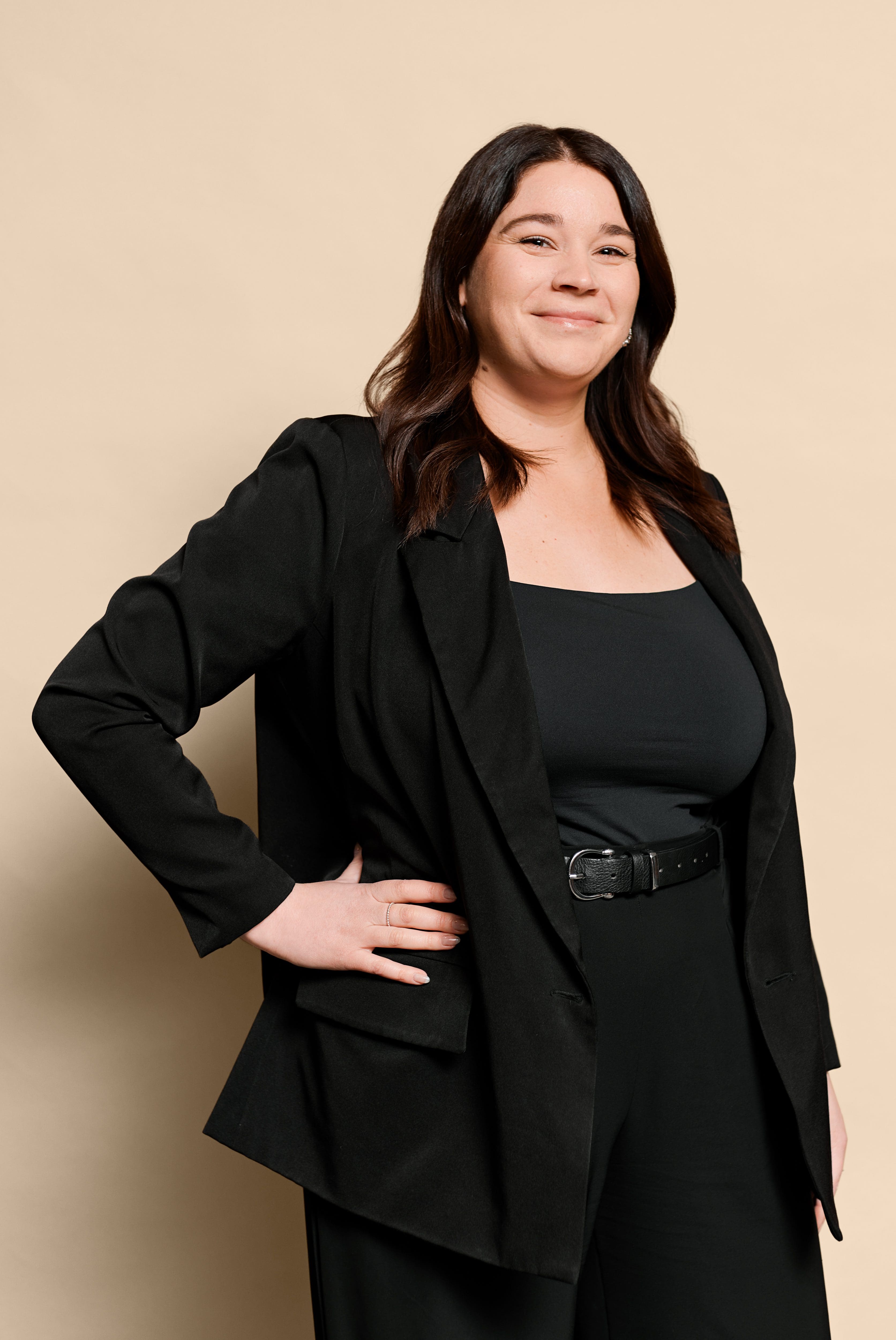 Une femme aux longs cheveux noirs, portant un blazer noir, un haut noir et une ceinture noire, se tient debout, souriante, une main sur la hanche, sur un fond beige à la Clinique médico-esthétique Viva.