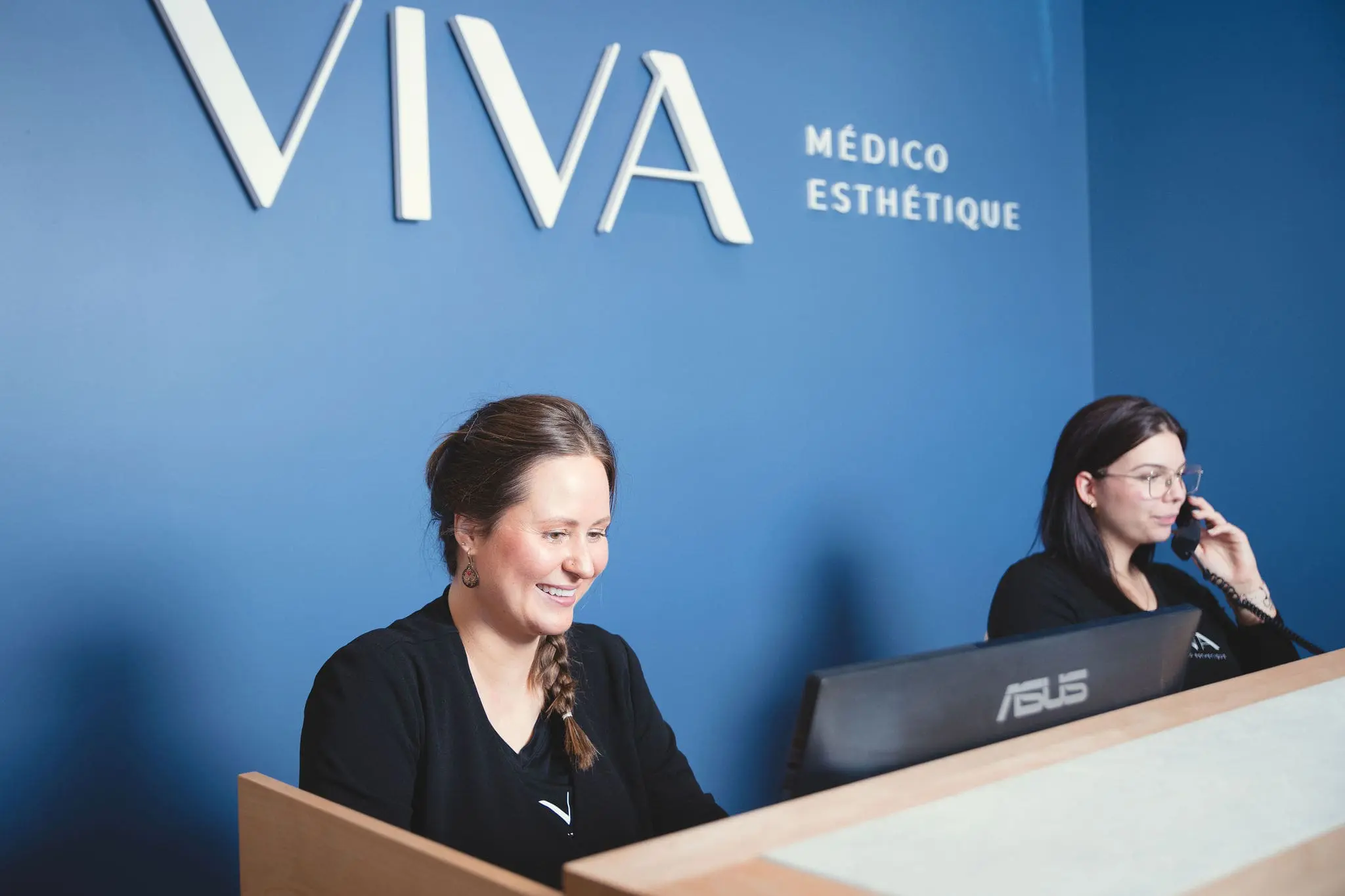 Deux femmes sont assises à la réception, devant un mur bleu où est écrit « VIVA MÉDICO ESTHÉTIQUE ». L'une sourit devant un ordinateur, tandis que l'autre parle au téléphone à la Clinique médico-esthétique Viva.