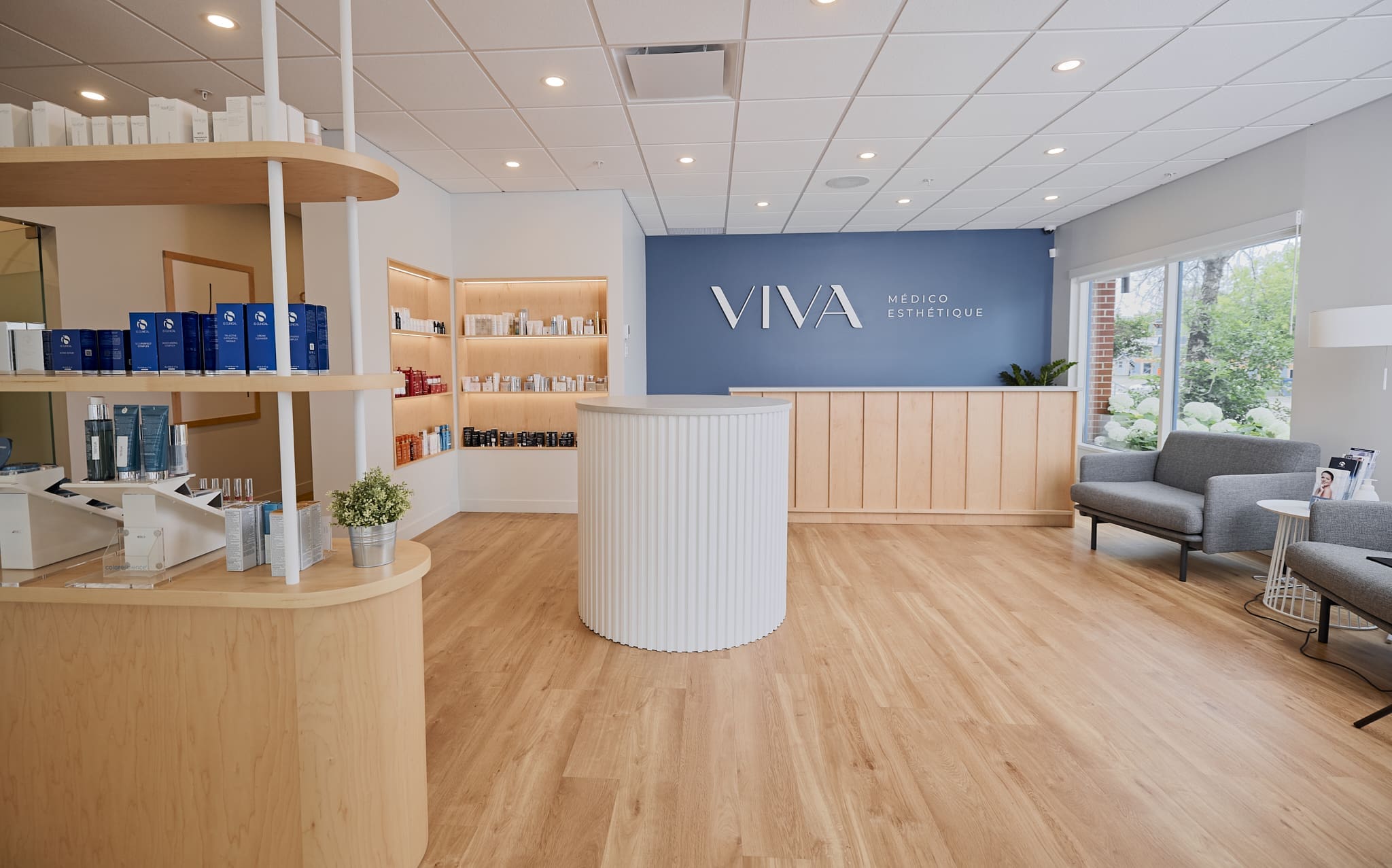 Une clinique d'esthétique médicale moderne et lumineuse avec des planchers de bois clair, un comptoir d'accueil blanc circulaire, des étagères de produits de soins de la peau, des chaises grises, de grandes fenêtres et un mur d'accent bleu avec la signalisation VIVA Médico Esthétique.