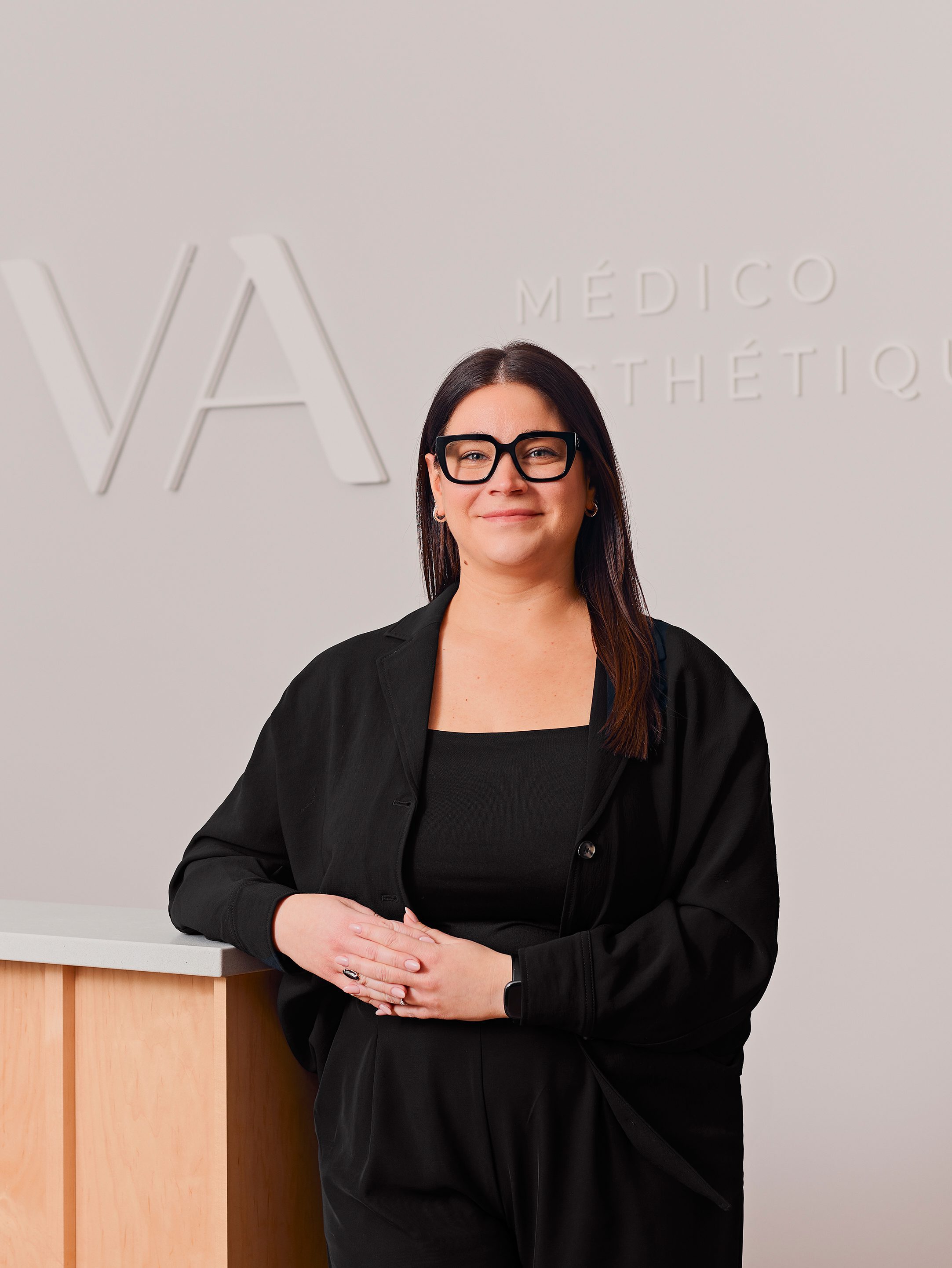 Une femme aux longs cheveux noirs et aux lunettes, vêtue de noir, se tient debout, souriante, devant un mur de couleur claire arborant une signalisation partielle MÉDICO ESTHÉTIQUE et de grandes initiales VA à la Clinique médico-esthétique Viva.