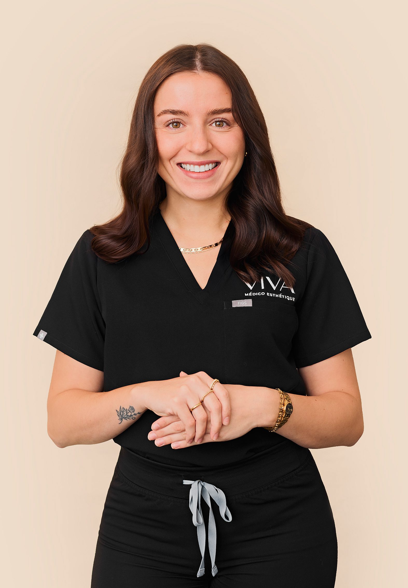 Une femme souriante aux cheveux bruns ondulés se tient debout sur un fond beige, vêtue d'une tenue médicale noire avec le logo Cliniques Viva Québec sur la poitrine. Elle porte un tatouage sur l'avant-bras, des bijoux en or et ses mains sont délicatement jointes.