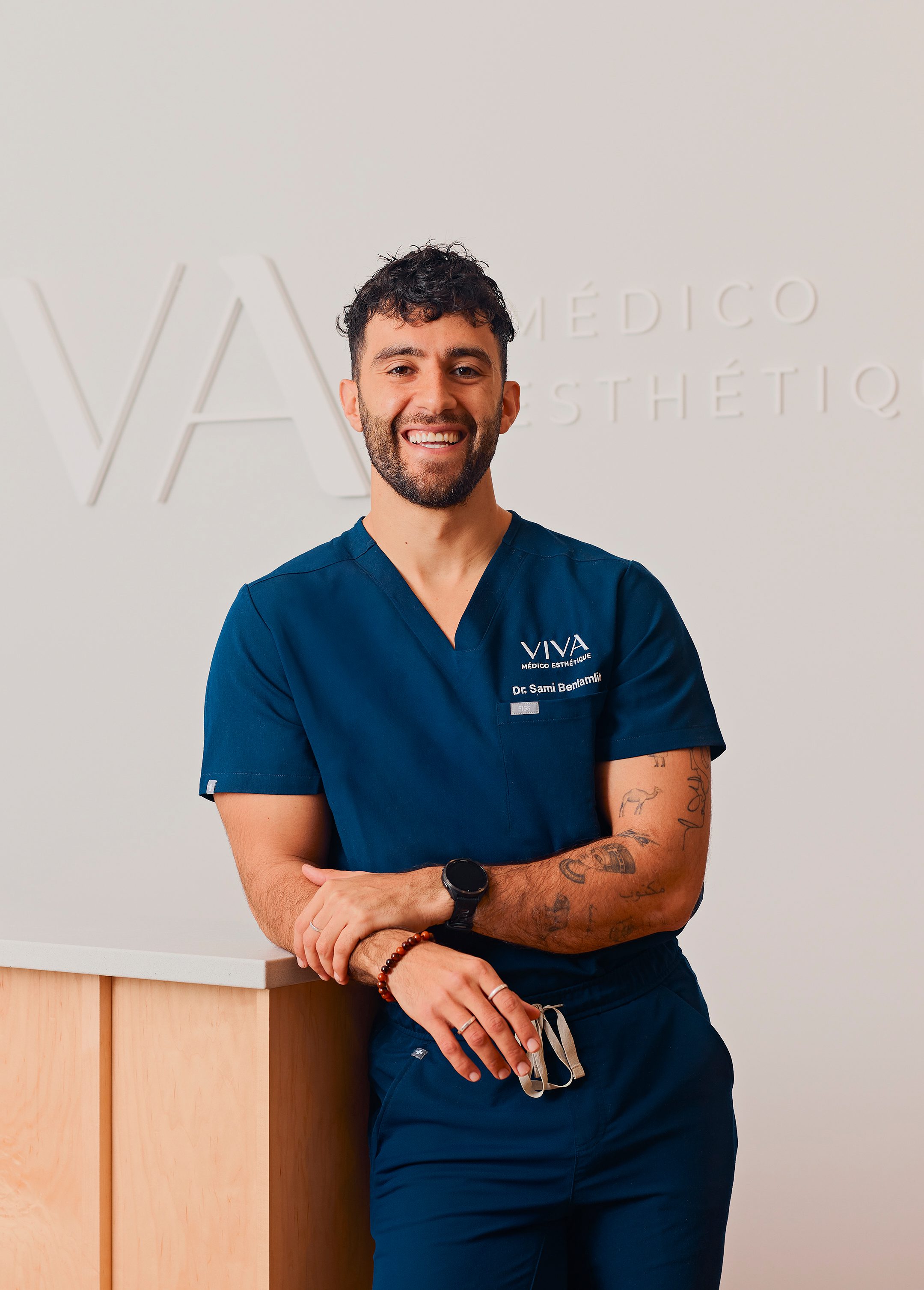 Un homme souriant, vêtu d'une tenue médicale bleu marine, se tient debout, les bras croisés, lunettes à la main. Il s'appuie contre un comptoir en bois dans un bureau moderne et minimaliste de la Clinique médico-esthétique Viva, avec le mot VIVA et une inscription discrète sur le mur derrière lui.
