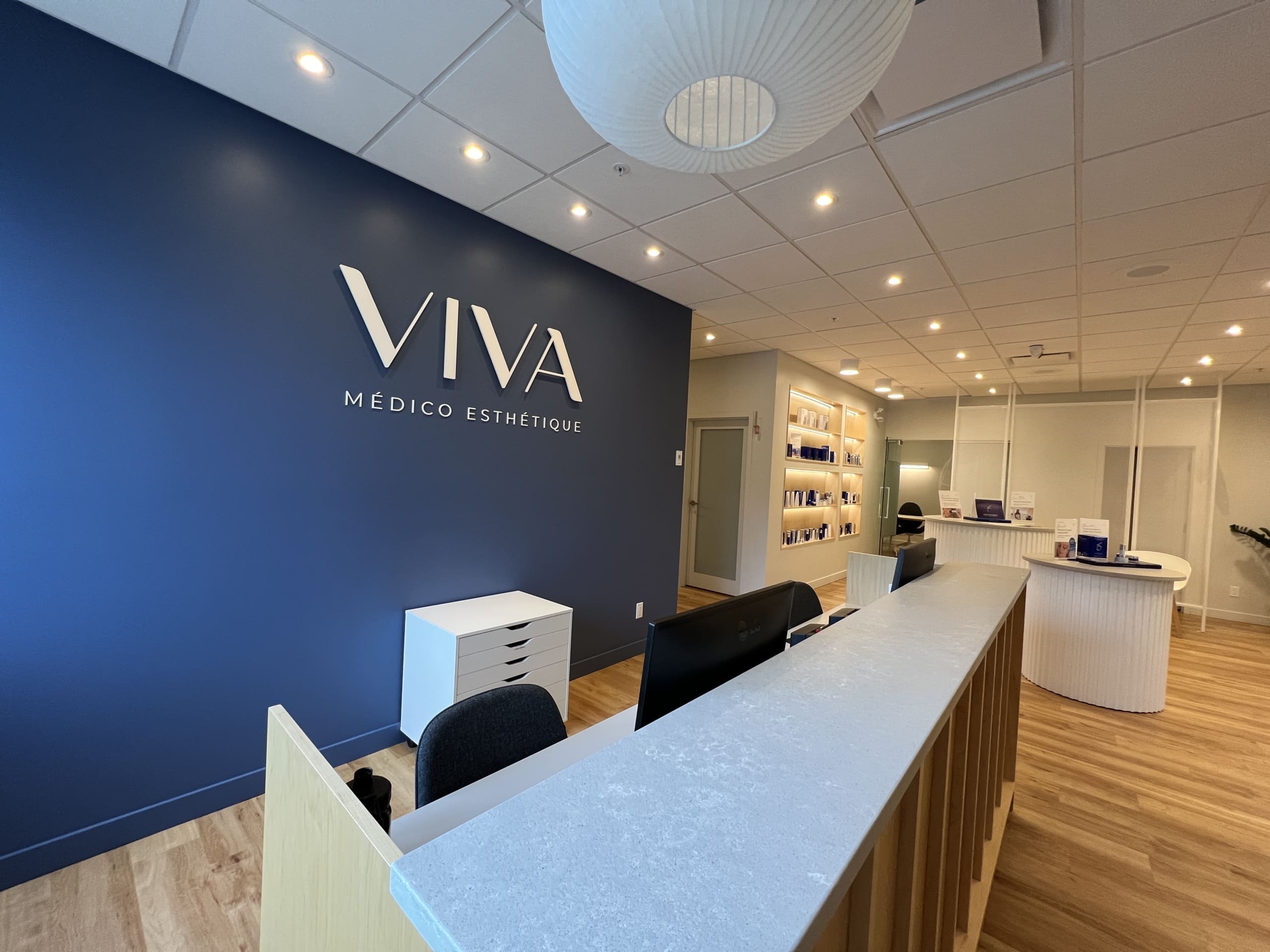 La réception moderne de VIVA Médico Esthétique avec un mur d'accent bleu, un bureau blanc élégant, des ordinateurs, un sol en bois et des étagères présentant des produits. L'espace est éclairé par des plafonniers.