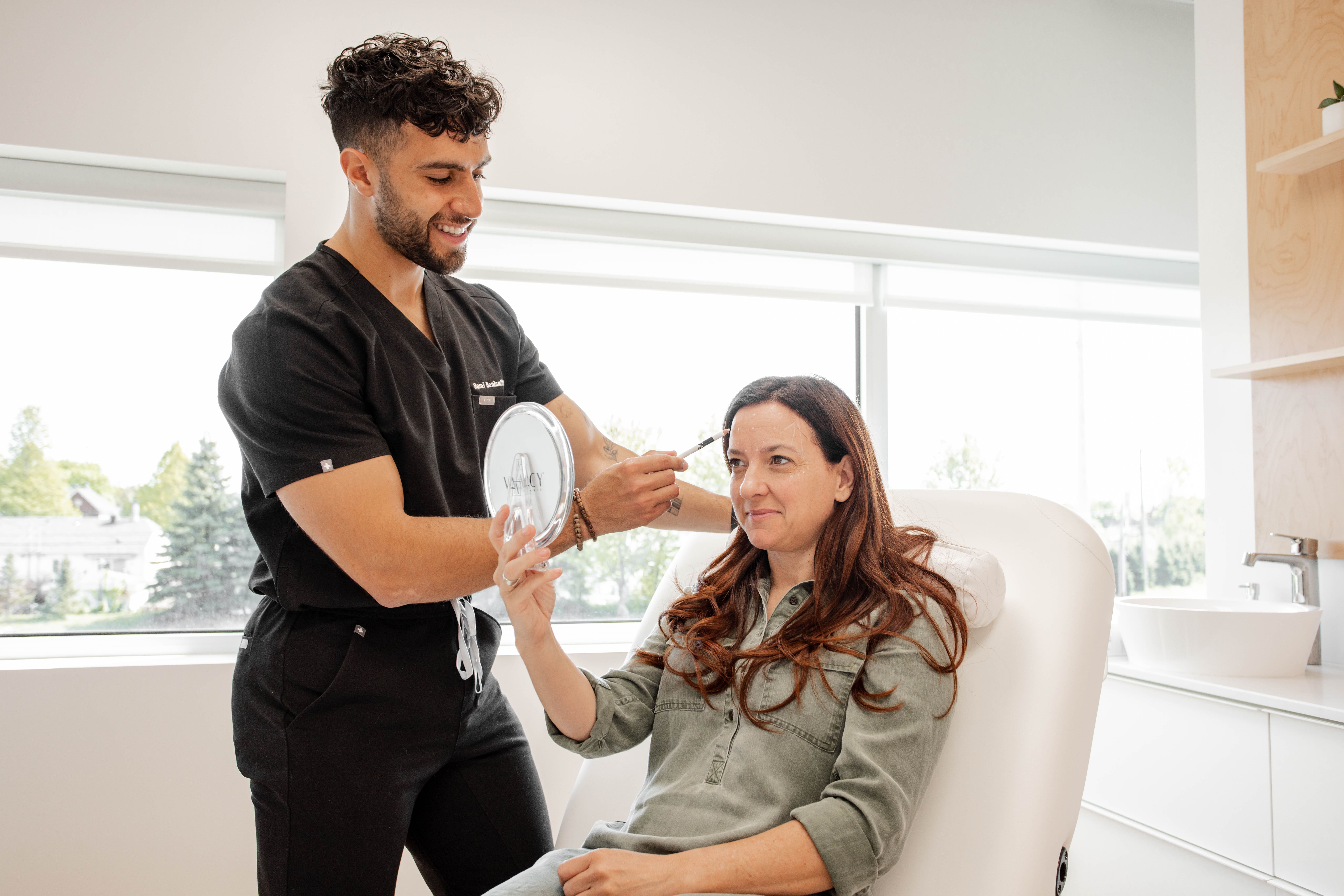 Un professionnel de la santé souriant en tenue noire se prépare à administrer une intervention esthétique à une femme assise tenant un petit miroir à la Clinique médico-esthétique Viva dans une salle de clinique lumineuse et moderne.