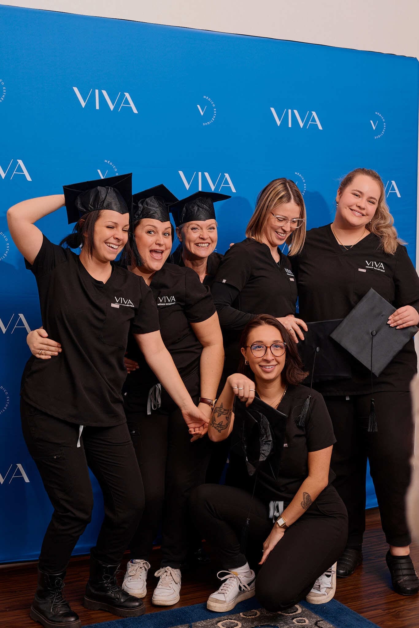 Six femmes en tenue noire et toque de diplômée posent et sourient devant un fond bleu portant l'inscription « VIVA », célébrant leur réussite à la Clinique médico-esthétique Viva. L'une est agenouillée devant, tenant sa toque, tandis que les autres se tiennent près les unes des autres.