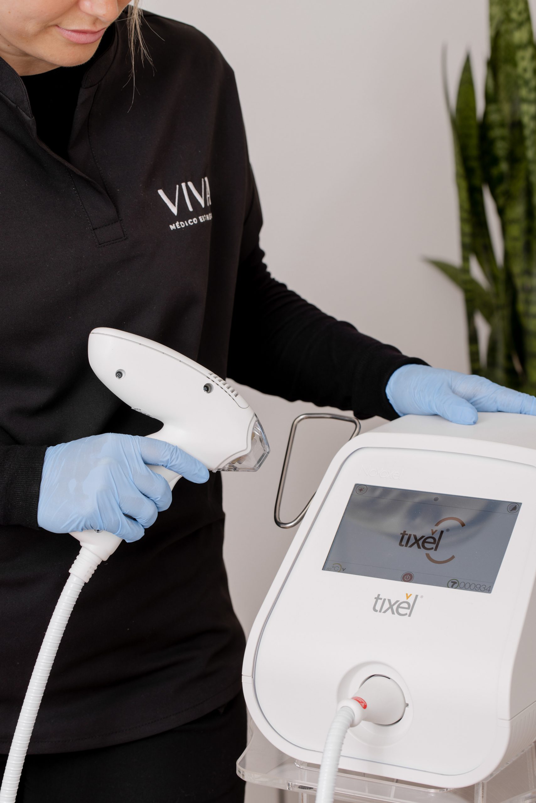 Une personne portant des gants bleus et une chemise noire tient un appareil portatif connecté à une machine Tixel avec un écran tactile, probablement dans la Clinique médico-esthétique Viva.