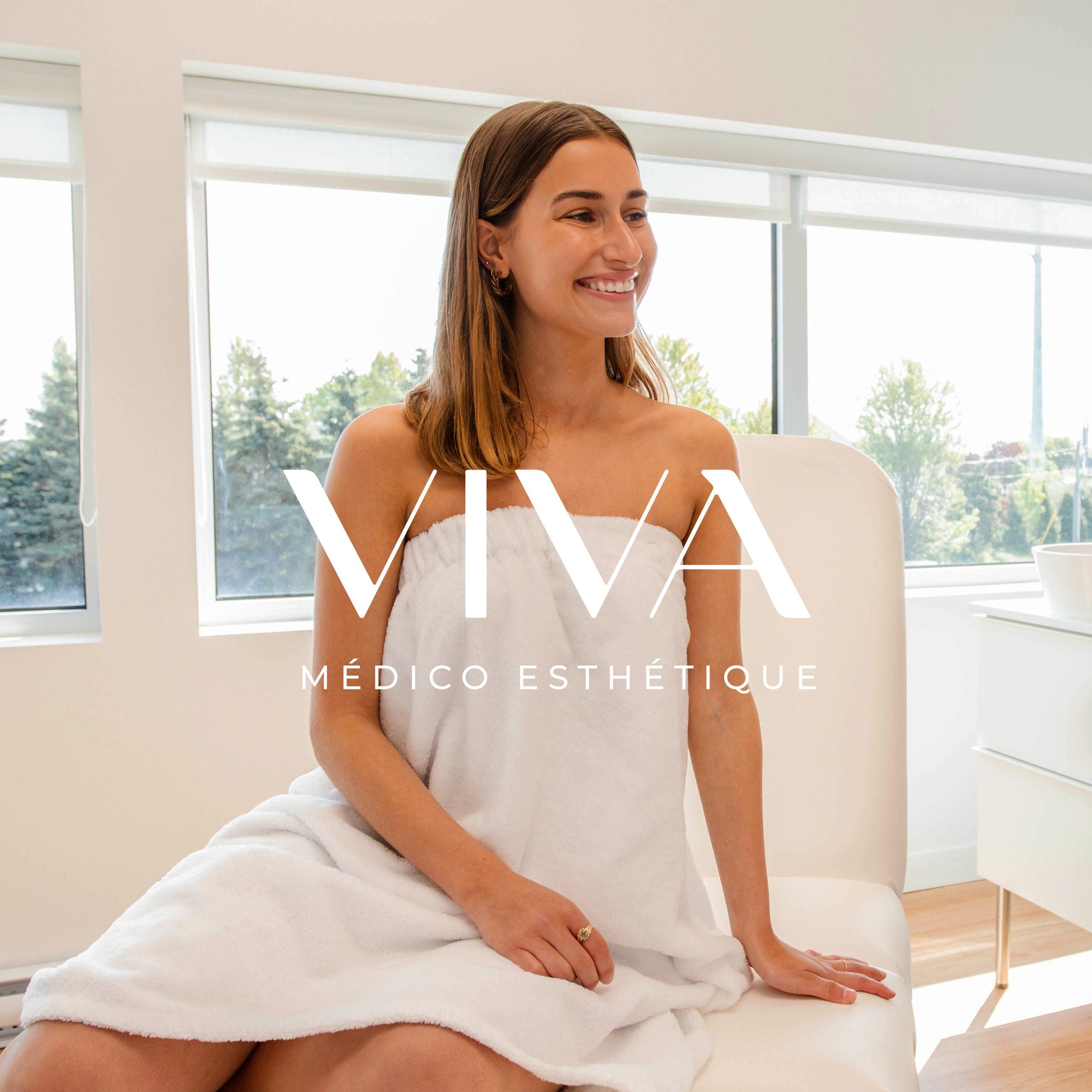 Une femme souriante, vêtue d'une serviette blanche, est assise dans une salle de consultation lumineuse et moderne. Le texte « VIVA MÉDICO ESTHÉTIQUE » est superposé à l'image. De grandes fenêtres laissent entrevoir les arbres et la lumière du soleil : découvrez l'ambiance sereine de la Clinique médico-esthétique Viva.