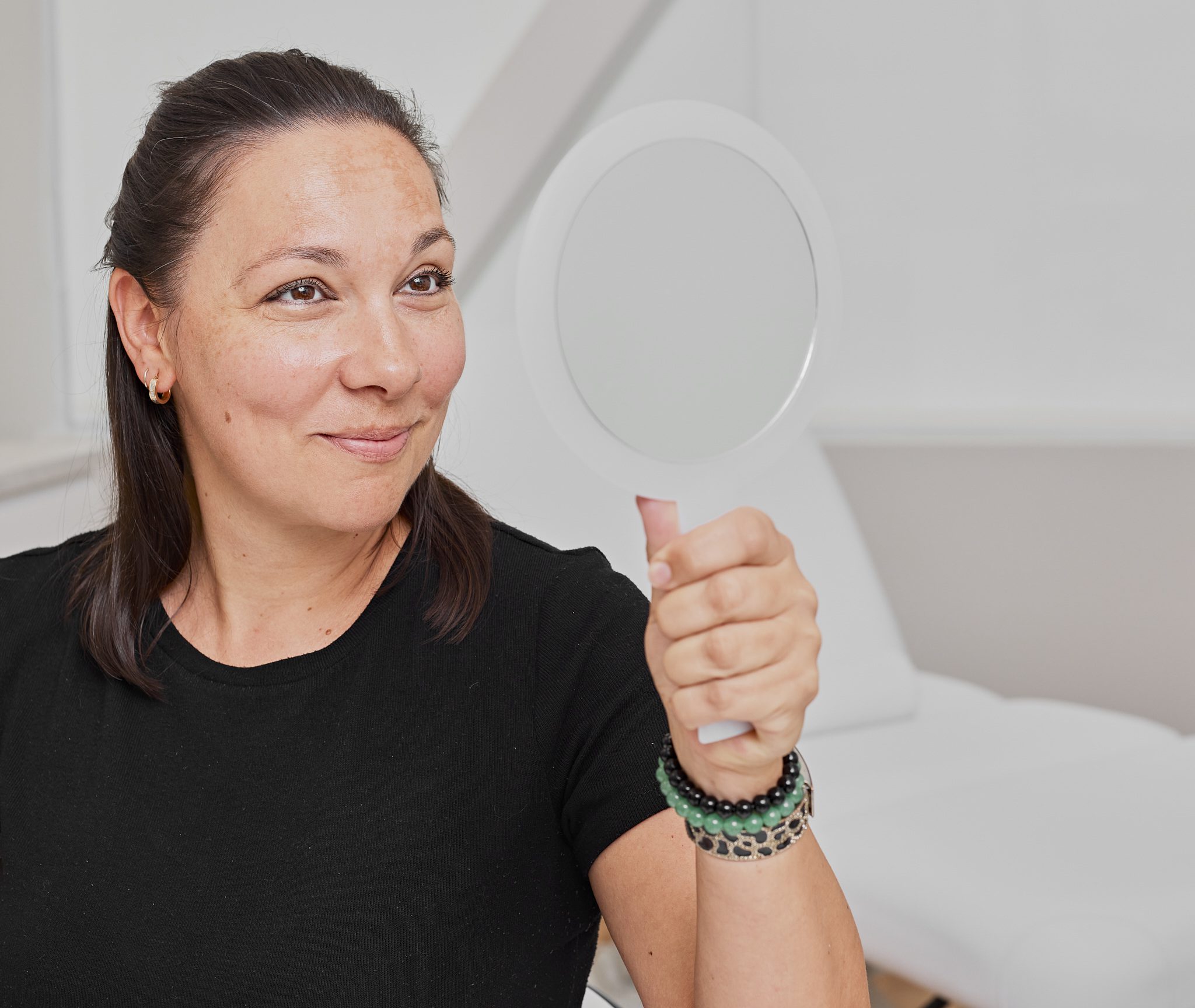 Une femme aux cheveux noirs, vêtue d'une chemise noire, sourit en se regardant dans un miroir à main. Elle porte un bracelet de perles et est assise dans une salle lumineuse et moderne de la Clinique médico-esthétique Viva.