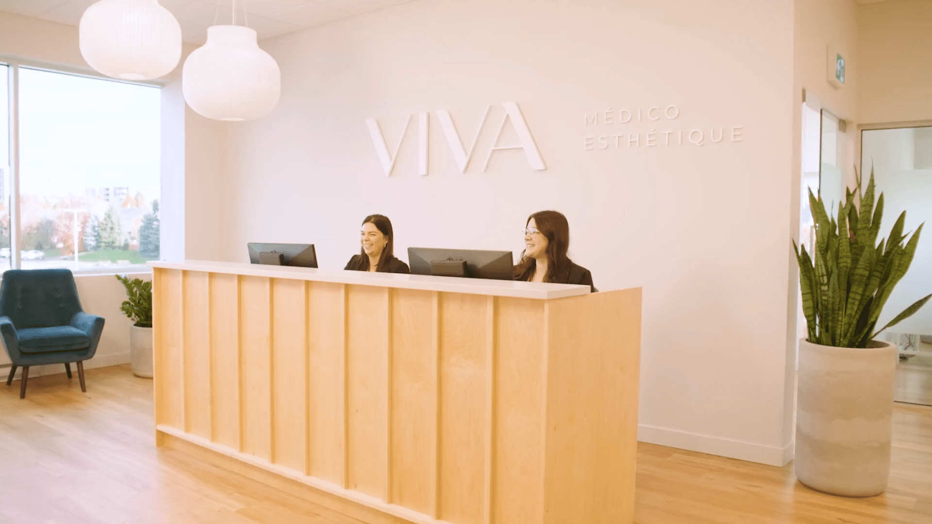 Deux femmes sont assises derrière un comptoir d'accueil en bois équipé d'ordinateurs à la Clinique médico-esthétique Viva. Le mur derrière elles affiche l'enseigne VIVA MÉDICO ESTHÉTIQUE, tandis que de grandes fenêtres et des plantes contribuent à la luminosité et à l'accueil chaleureux de l'espace.