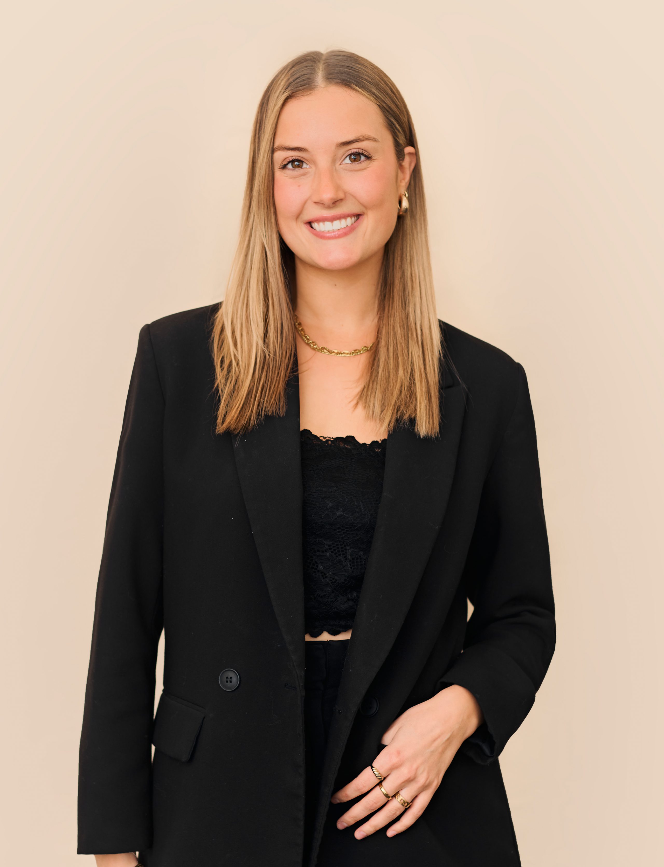 Une femme aux cheveux blonds raides et mi-longs sourit à l'objectif. Elle porte un blazer noir sur un haut en dentelle noire, des bijoux en or et se tient debout sur un fond beige à la Clinique médico-esthétique Viva.