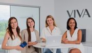 Quatre femmes sont assises à une table blanche dans un bureau moderne près d'une fenêtre, avec un ordinateur portable et un produit Clinique médico-esthétique Viva. Elles sourient, le mot VIVA étant affiché sur le mur derrière elles.