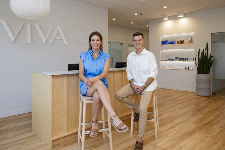 Deux personnes, une femme en robe bleue et un homme en chemise blanche et pantalon beige, sont assis sur des tabourets dans le hall moderne et lumineux de la Clinique médico-esthétique Viva, avec une enseigne « VIVA » et des étagères avec des produits en arrière-plan.