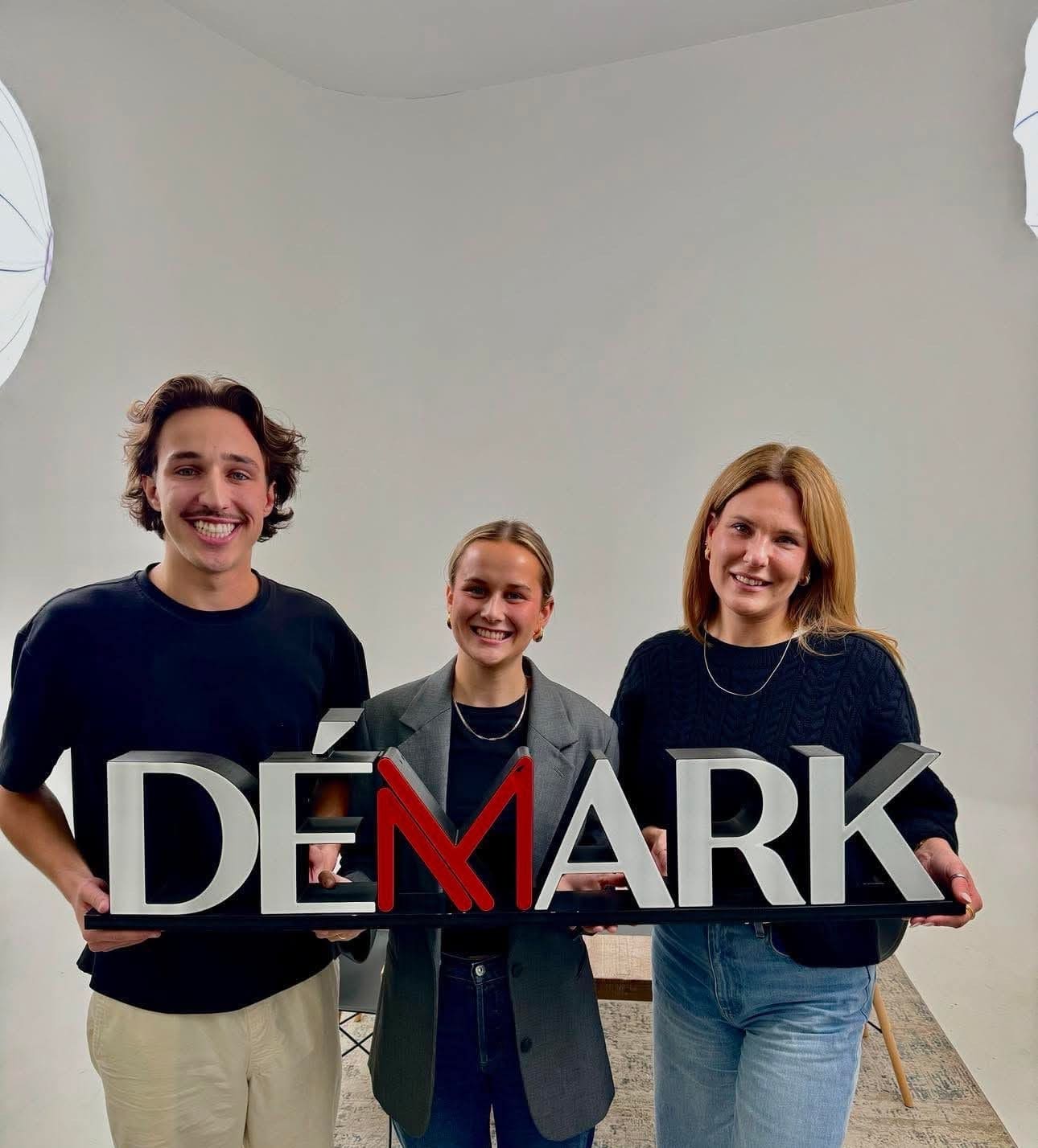 Trois personnes se tiennent à l'intérieur, tenant une grande pancarte portant l'inscription DÉMARK. Elles sourient et se tiennent devant un fond blanc uni, sous un éclairage tamisé, représentant les Cliniques Viva Québec et l'esprit de la Clinique médico-esthétique Viva.