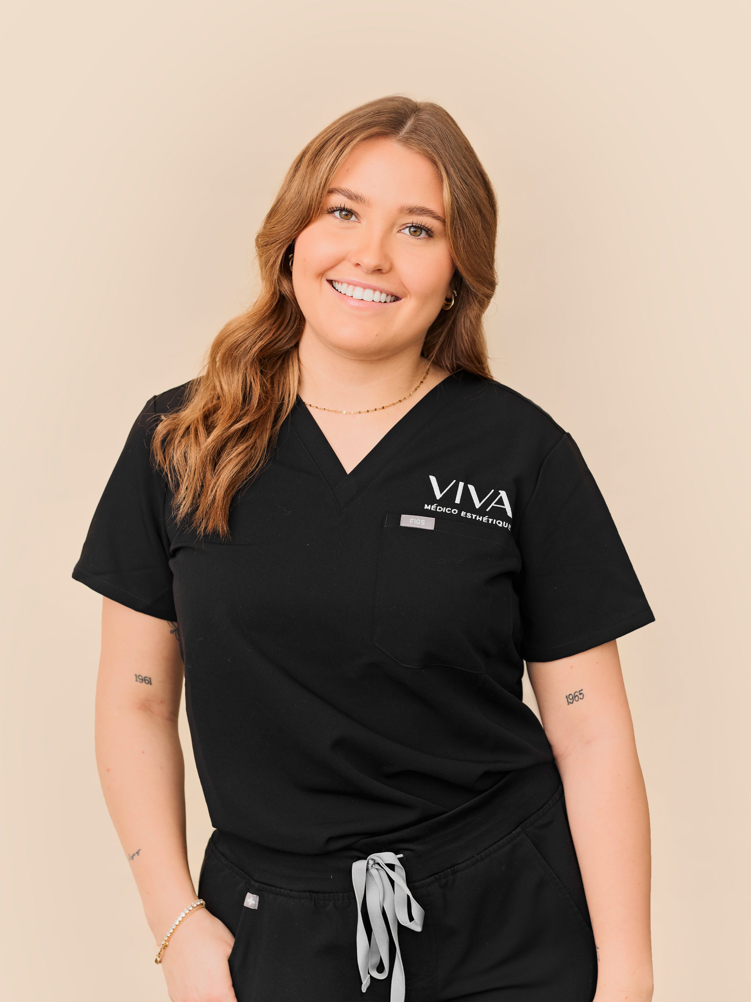 Une femme souriante aux cheveux châtain clair ondulés portant une blouse médicale noire avec l'inscription Clinique médico-esthétique Viva écrite sur le dessus se dresse sur un fond beige clair.