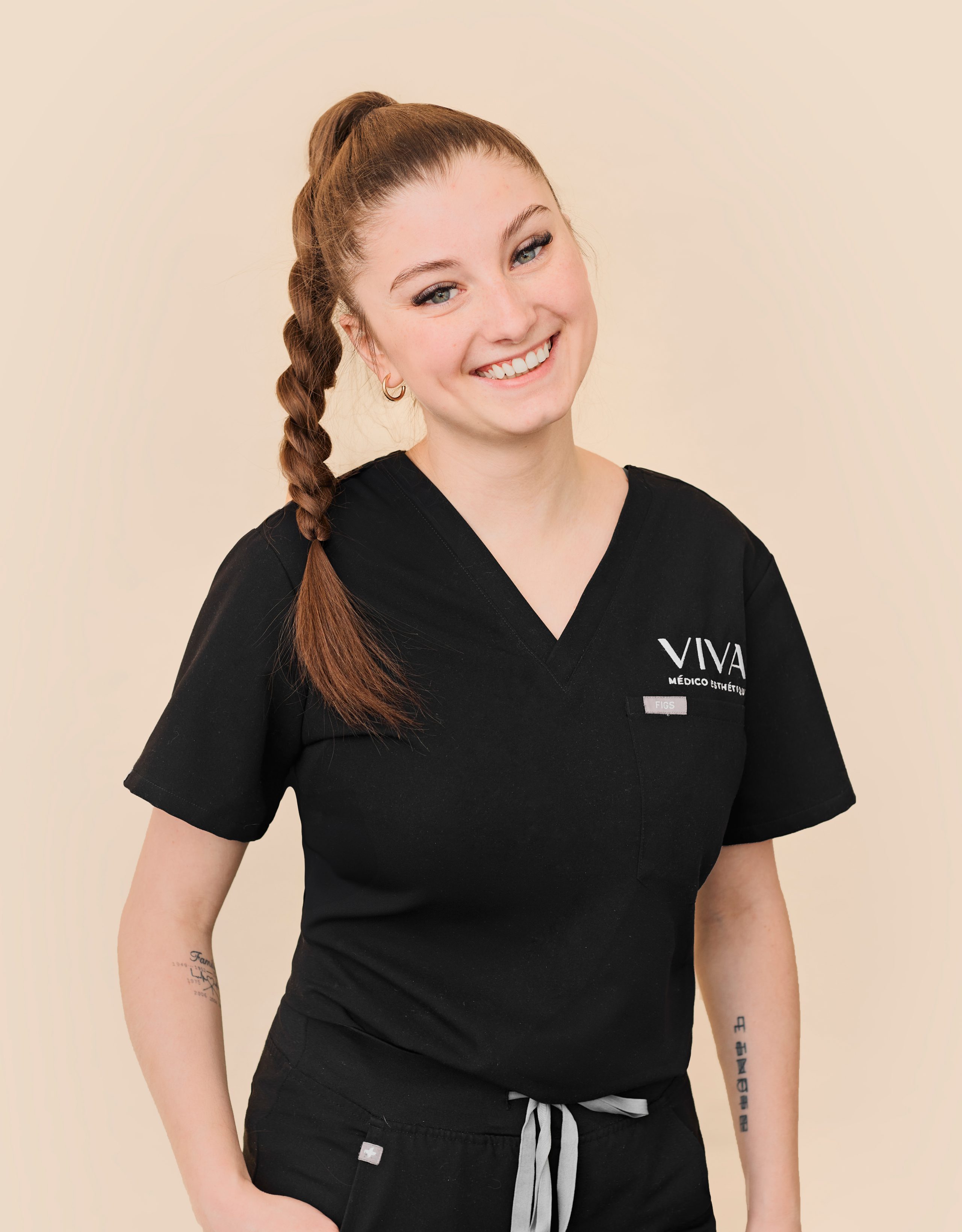 Une femme souriante avec une longue queue de cheval tressée, vêtue d'une blouse médicale noire avec le logo des Cliniques Viva Québec sur la poitrine, se tient debout sur un fond beige uni.