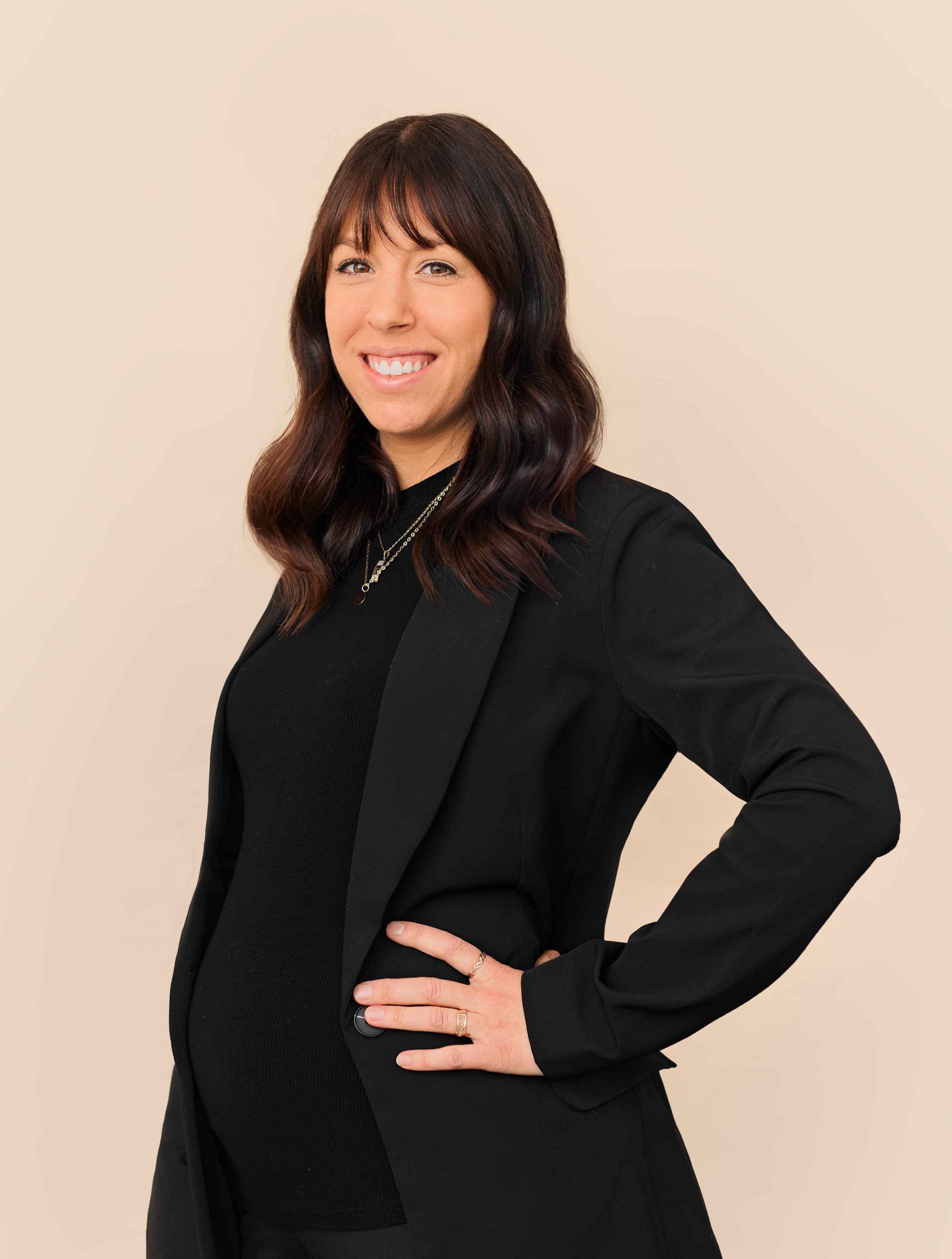 Une femme aux longs cheveux bruns, vêtue d'un blazer noir et d'un haut noir, se tient debout de côté, une main sur la hanche, souriant devant un fond beige uni à la Clinique médico-esthétique Viva.