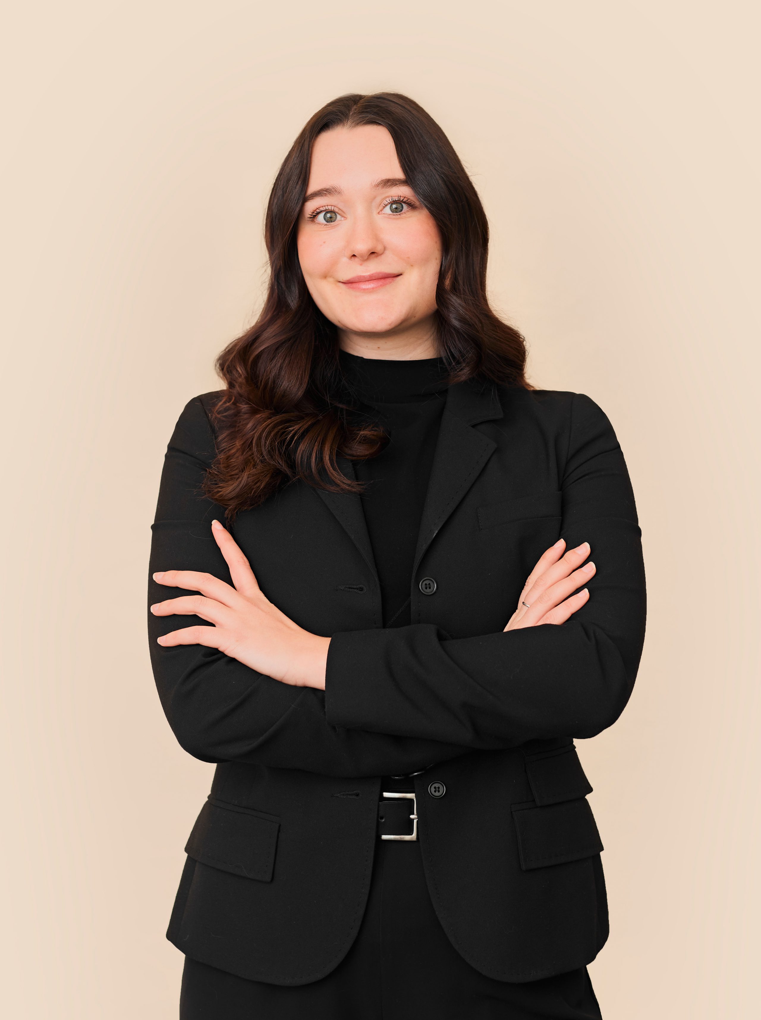 Une femme aux longs cheveux bruns vêtue d'un costume noir se tient debout, les bras croisés, regardant avec assurance la caméra sur un fond beige, incarnant le professionnalisme de la Clinique médico-esthétique Viva.