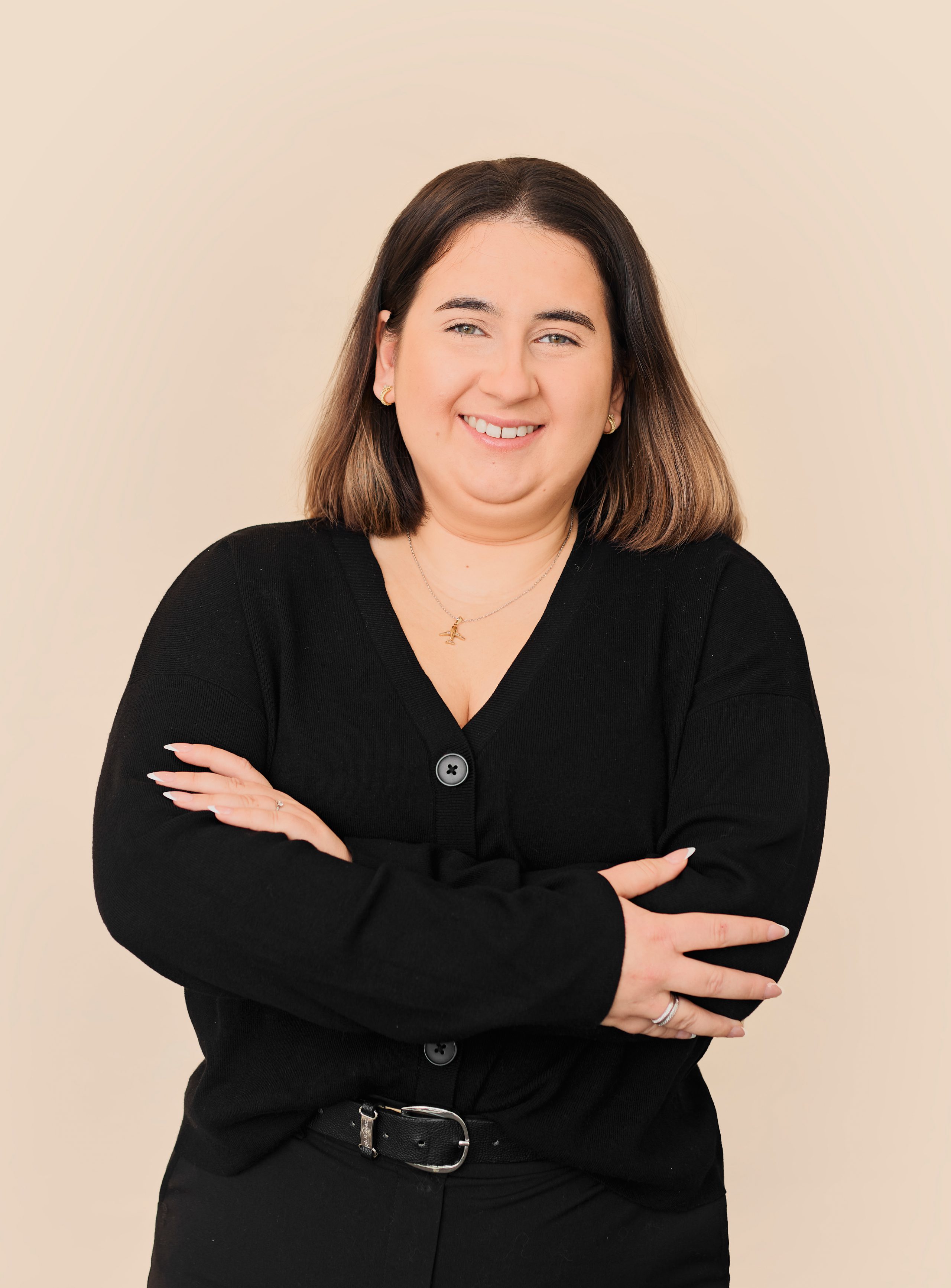 Une femme aux cheveux bruns et raides sourit avec assurance, les bras croisés, vêtue d'un cardigan noir, d'un pantalon noir et d'un collier en or. Elle se tient devant un fond beige, représentant l'équipe d'accueil de la Clinique médico-esthétique Viva.
