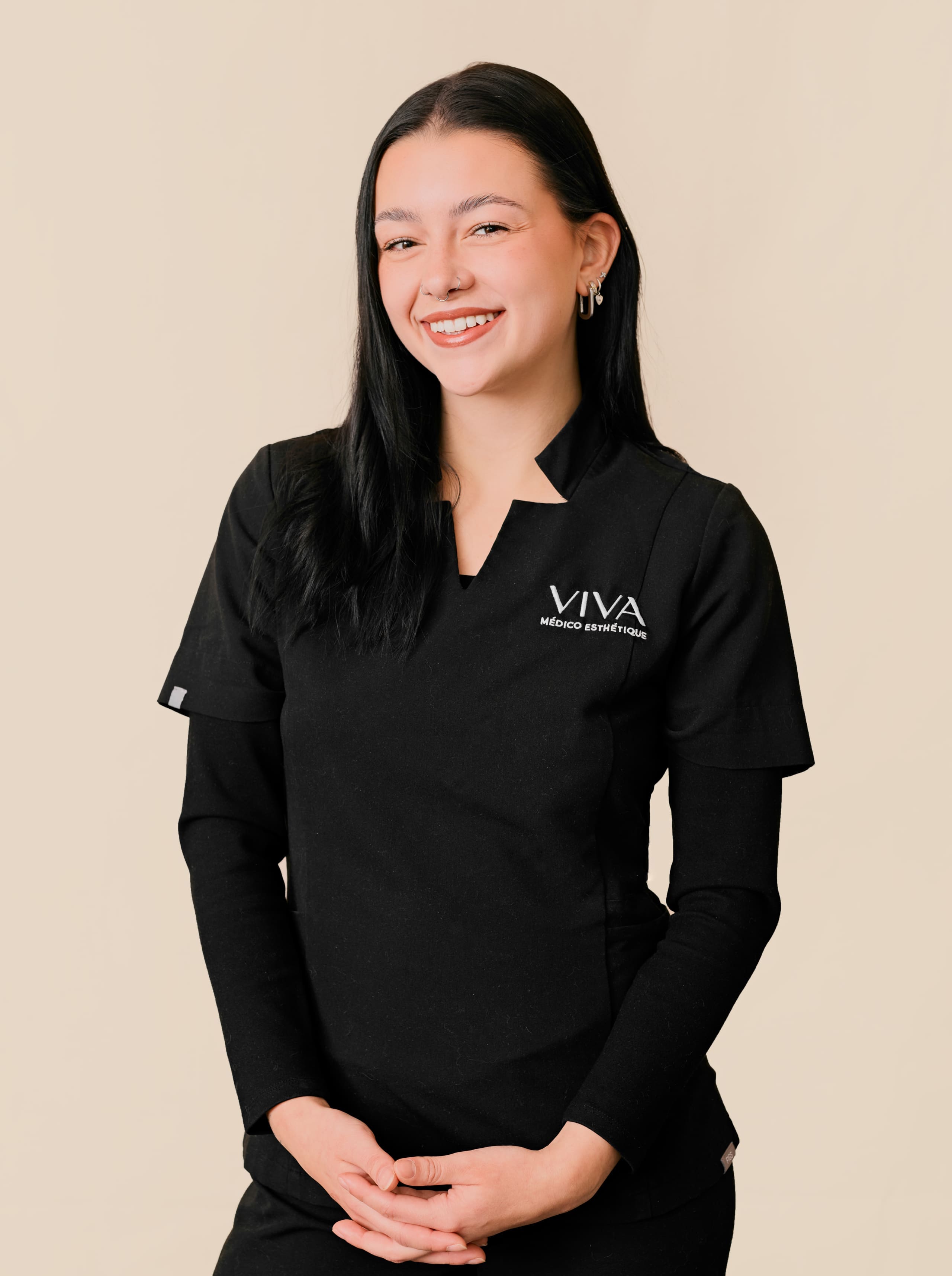 Une femme aux longs cheveux bruns, portant l'uniforme noir de VIVA Medical Esthetics, se tient sur un fond beige, souriante, les mains délicatement jointes devant elle.