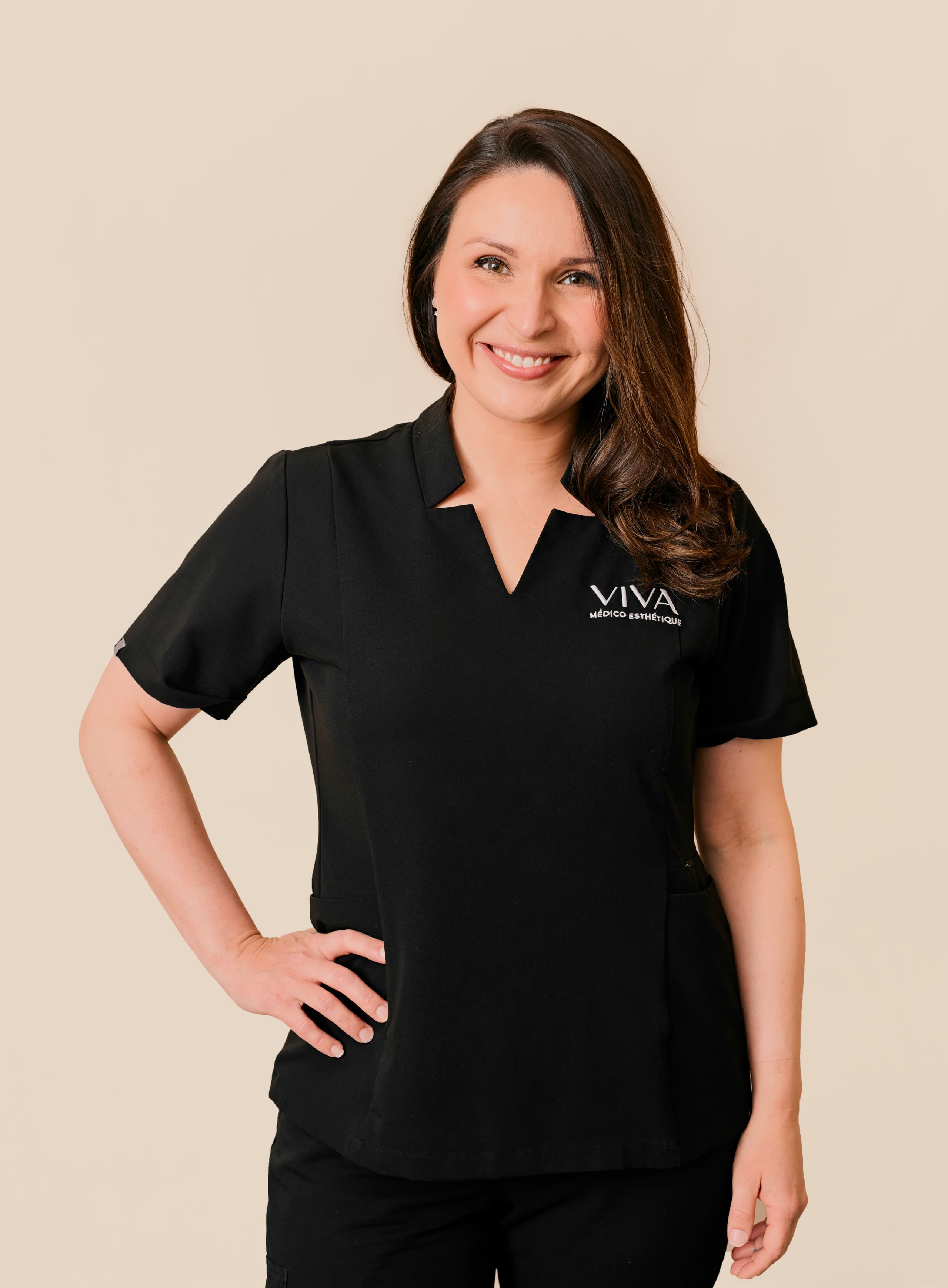Une femme aux longs cheveux bruns portant un uniforme noir avec le logo VIVA ultimate medspa sourit sur un fond beige clair, une main sur la hanche.