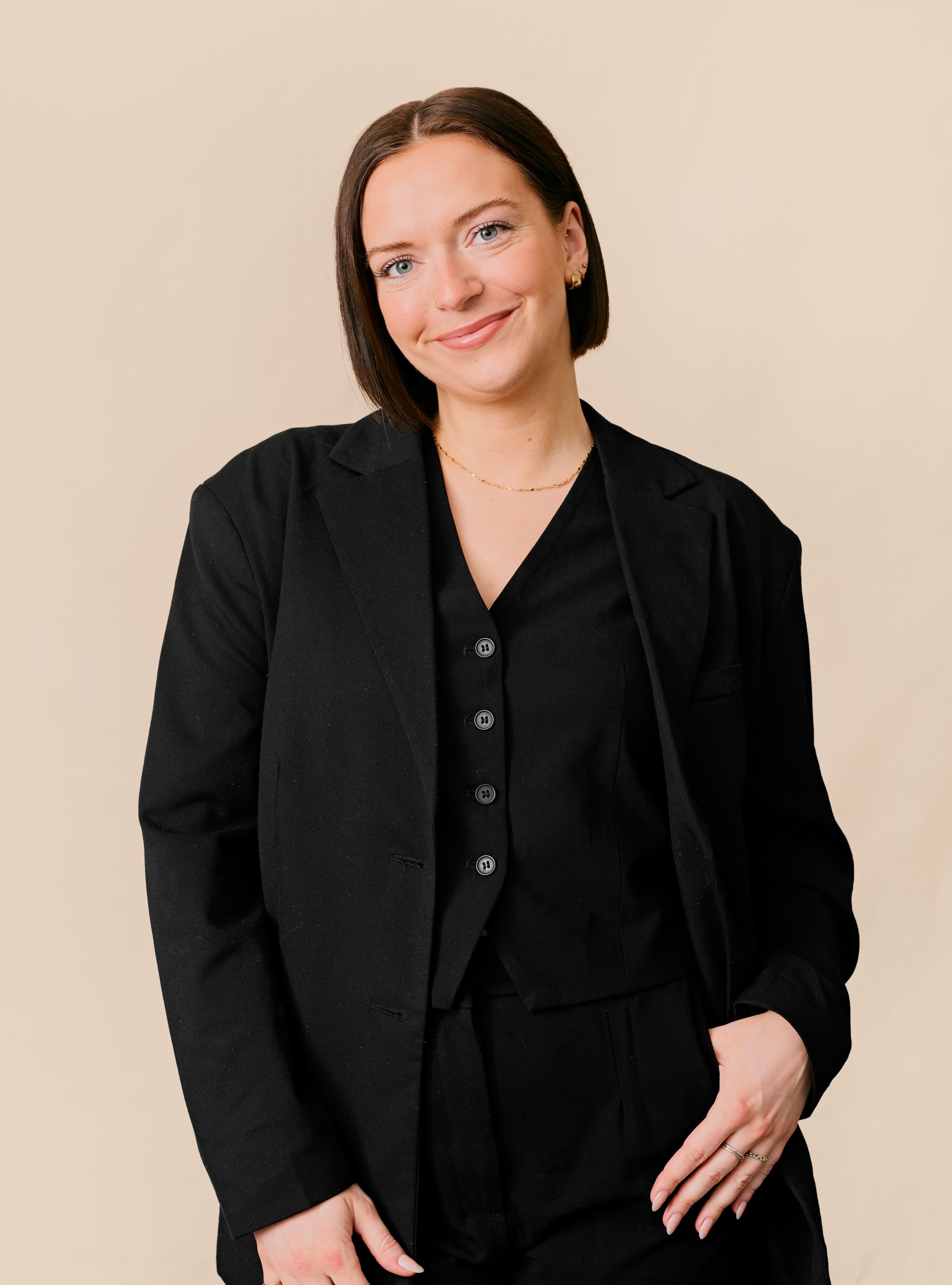 Une femme aux cheveux bruns raides, portant un blazer noir et un gilet assorti, sourit sur un fond beige uni.