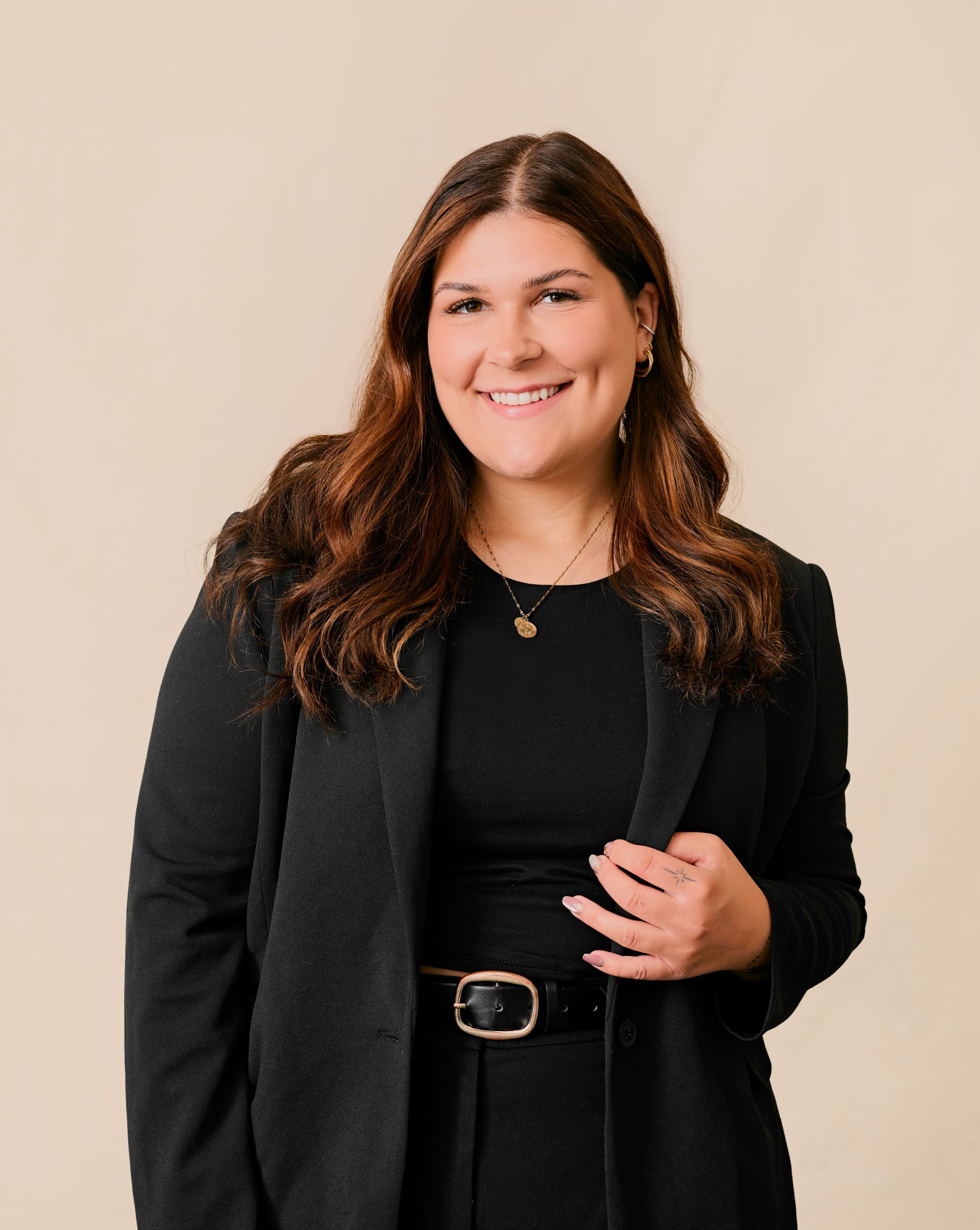 Une femme aux longs cheveux bruns portant un blazer noir, un haut noir et un collier doré sourit à l'appareil photo sur un fond beige neutre. Elle tient d'une main le revers de son blazer.